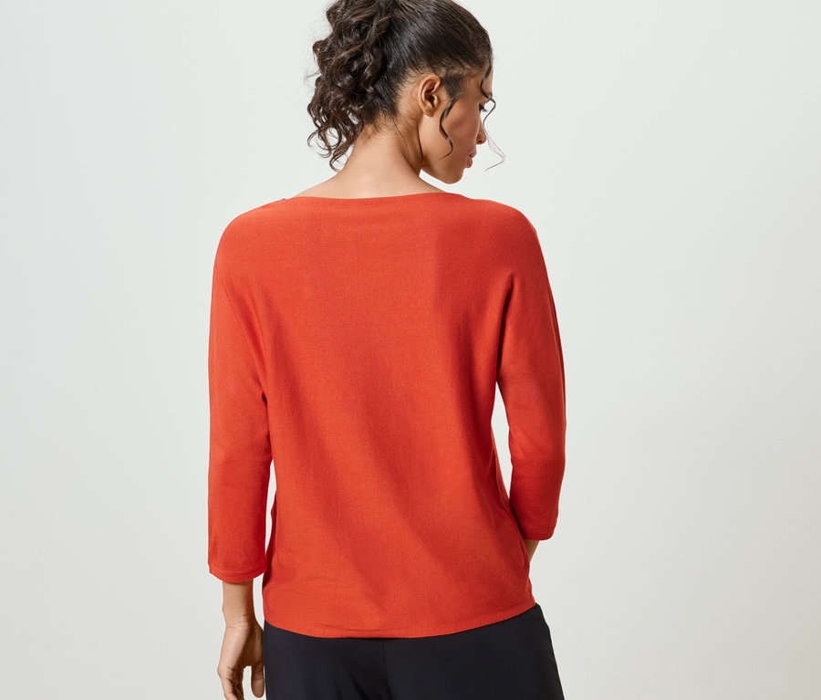 Zozadu je vidieť žena s vlasmi stiahnutými do copu, má na sebe oranžový pulóver z jemného úpletu a čierne nohavice.