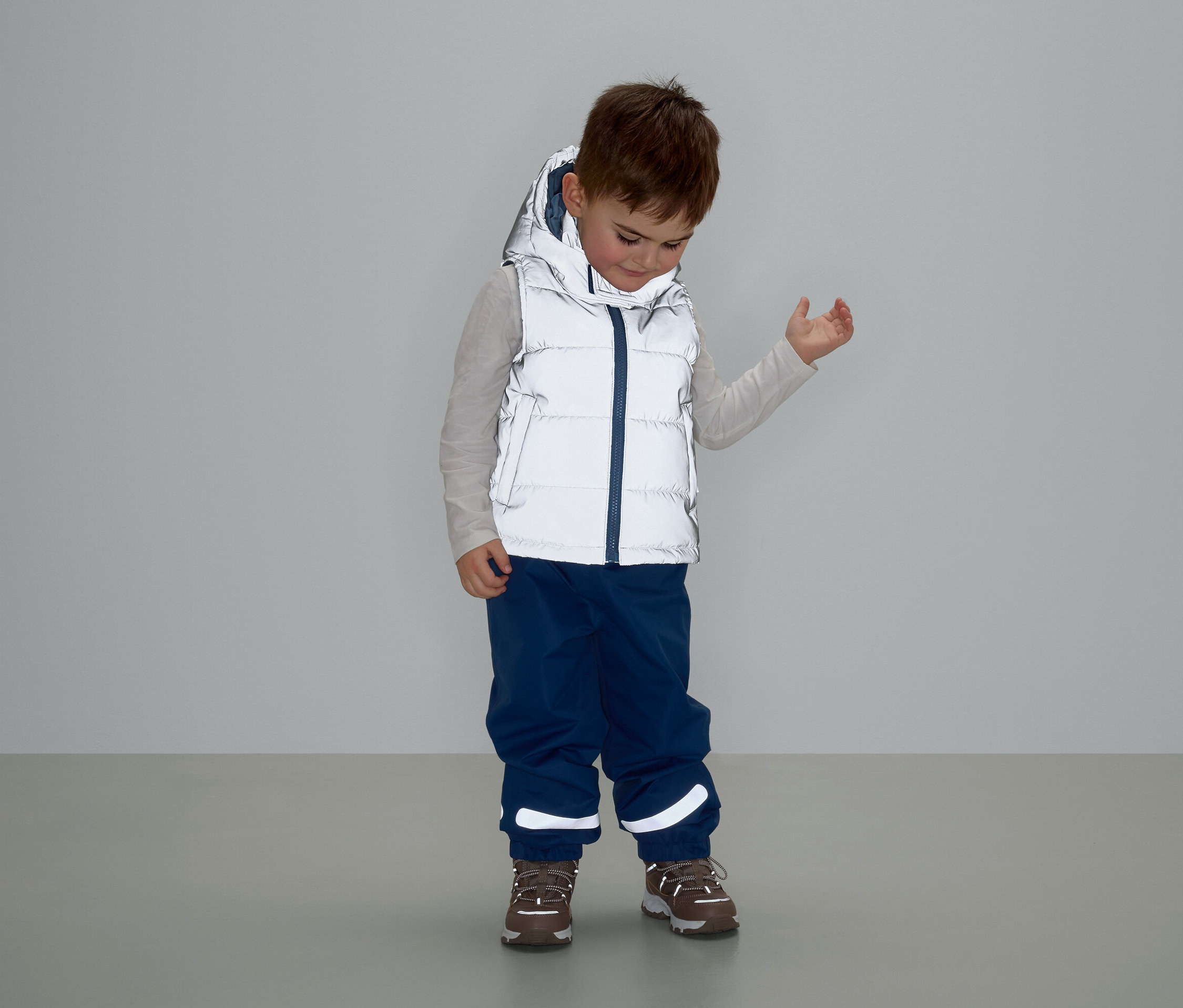 Chlapec v šedej veste s kapucňou, modrých nohaviciach a hnedých topánkach stojí a pozerá sa dole.