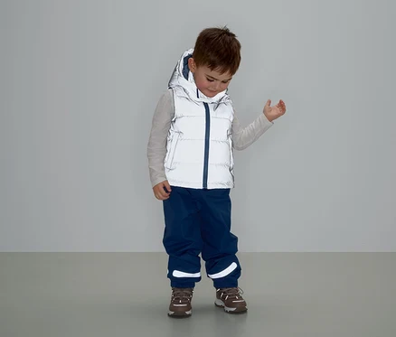 Chlapec v šedej veste s kapucňou, modrých nohaviciach a hnedých topánkach stojí a pozerá sa dole.