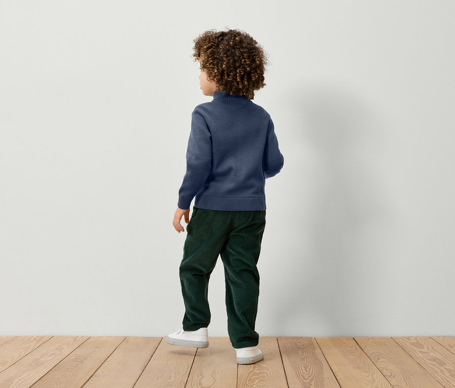 Chlapec s kučeravými vlasmi stojí chrbtom a má na sebe modrý sveter, zelené nohavice a biele tenisky.