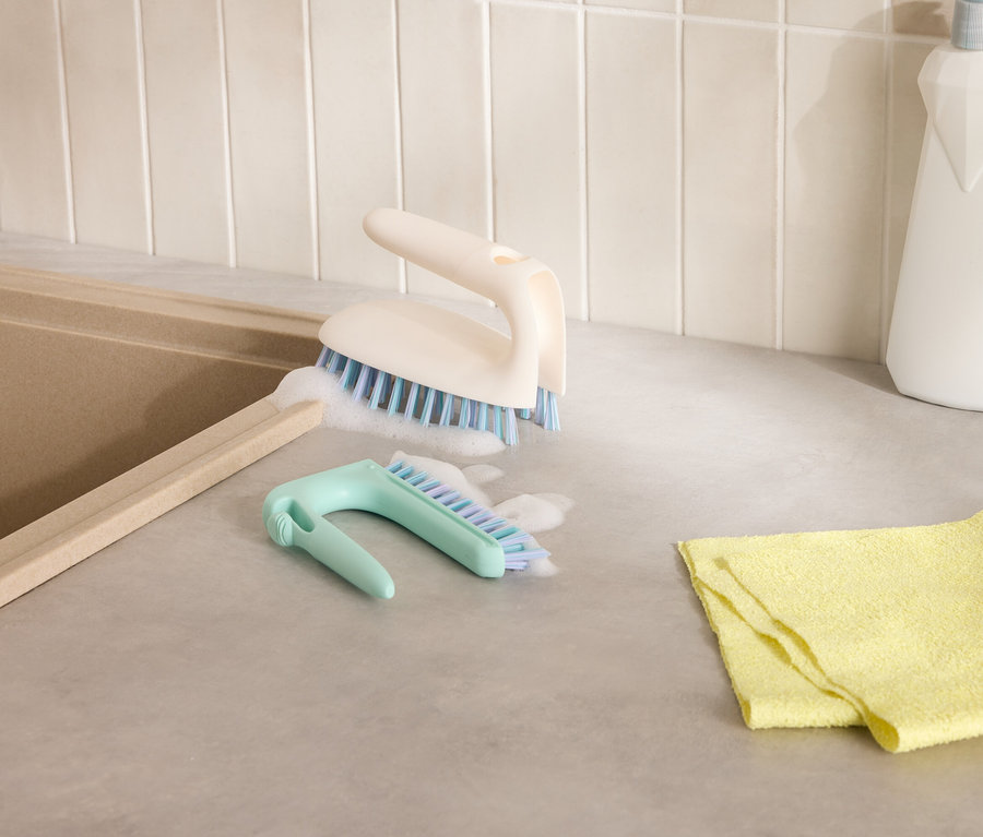 Dve čistiace kefy a žltá handrička na sivej doske stola vedľa drezu.