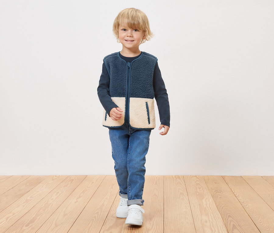 Chlapec v tmavomodrej flísovej veste s krémovými spodnými panelmi na zips stojí na drevenej podlahe.