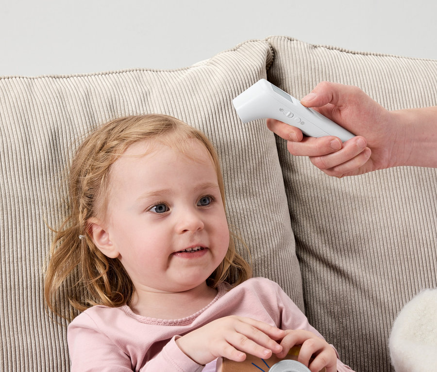 Malé dievča leží na pohovke, zatiaľ čo ruka drží biely digitálny teplomer pri jej čele.