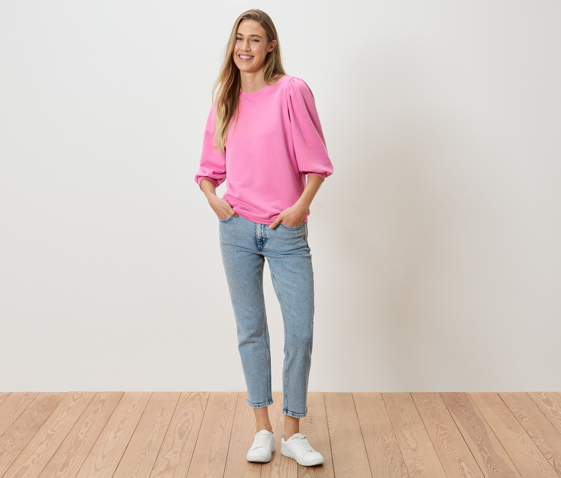 Žena s rukami vo vreckách nohavíc má na sebe ružový top s balónovými rukávmi, džínsy a biele tenisky.