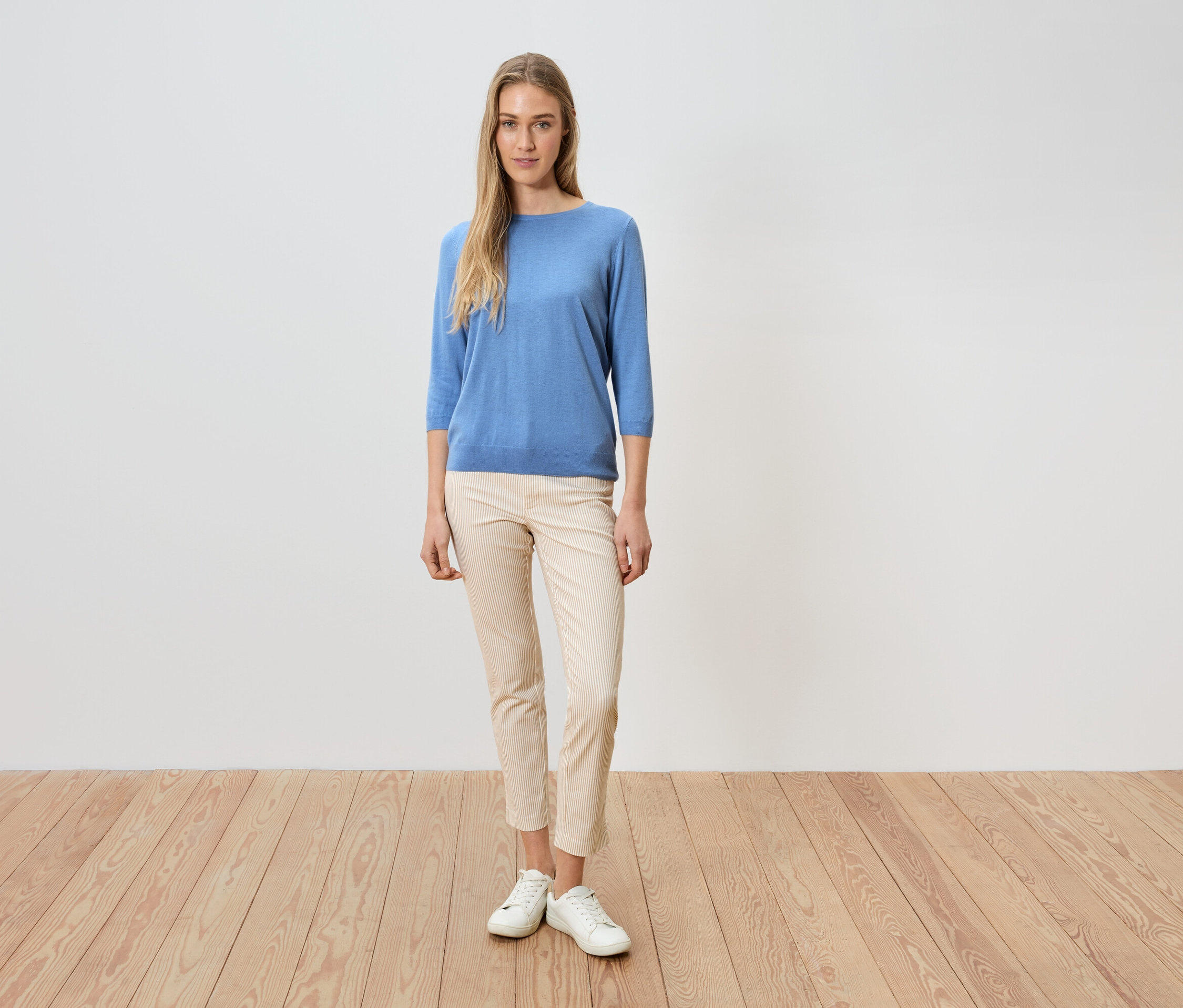 Žena stojí pred bielou stenou, má na sebe modrý sveter, svetlé nohavice a biele topánky.