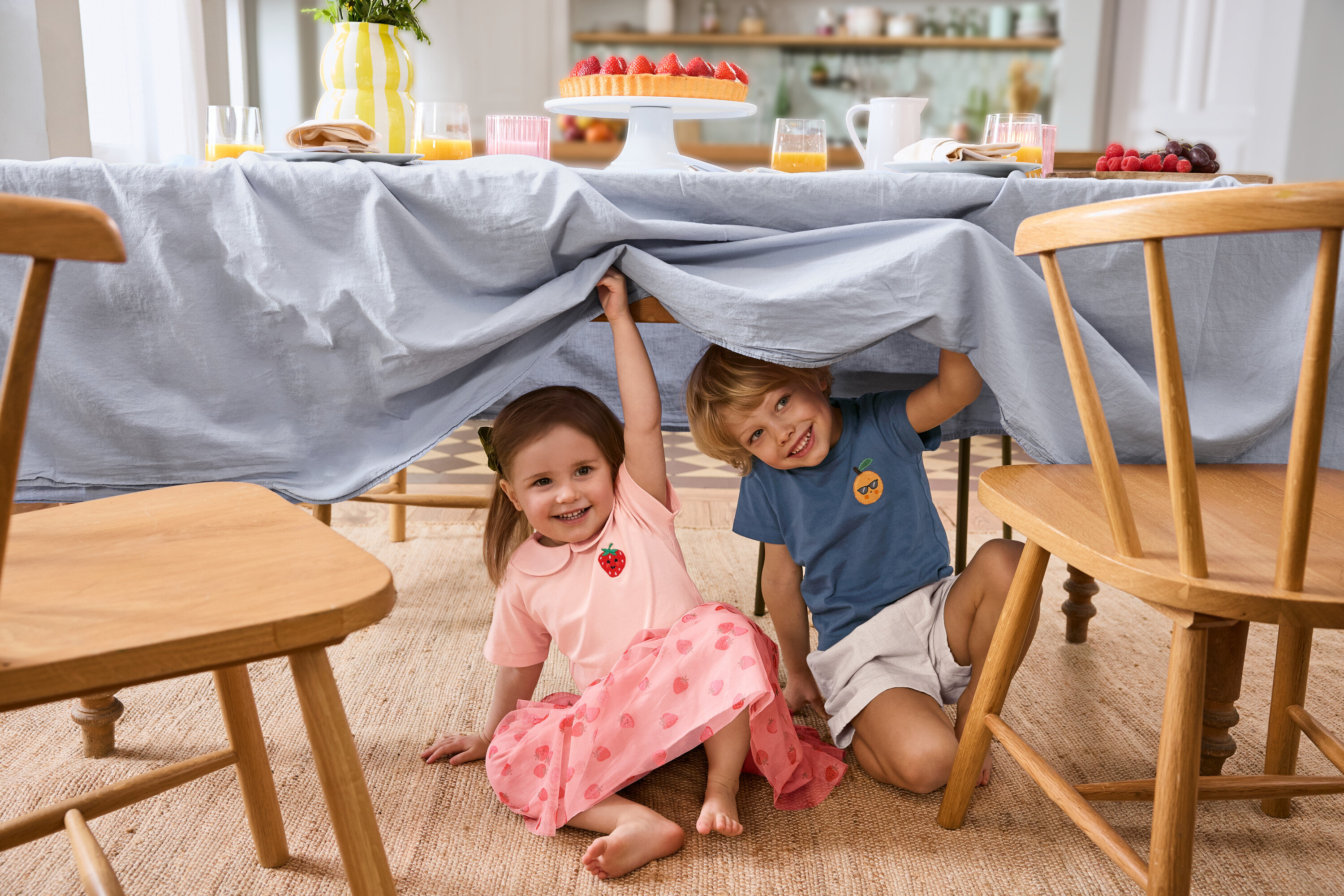 Dve deti sedia pod stolom s bledomodrým obrusom. Dievča má na sebe Detskú tylovú sukňu a 2 Detské tričká. Chlapec má na sebe Detské šortky.