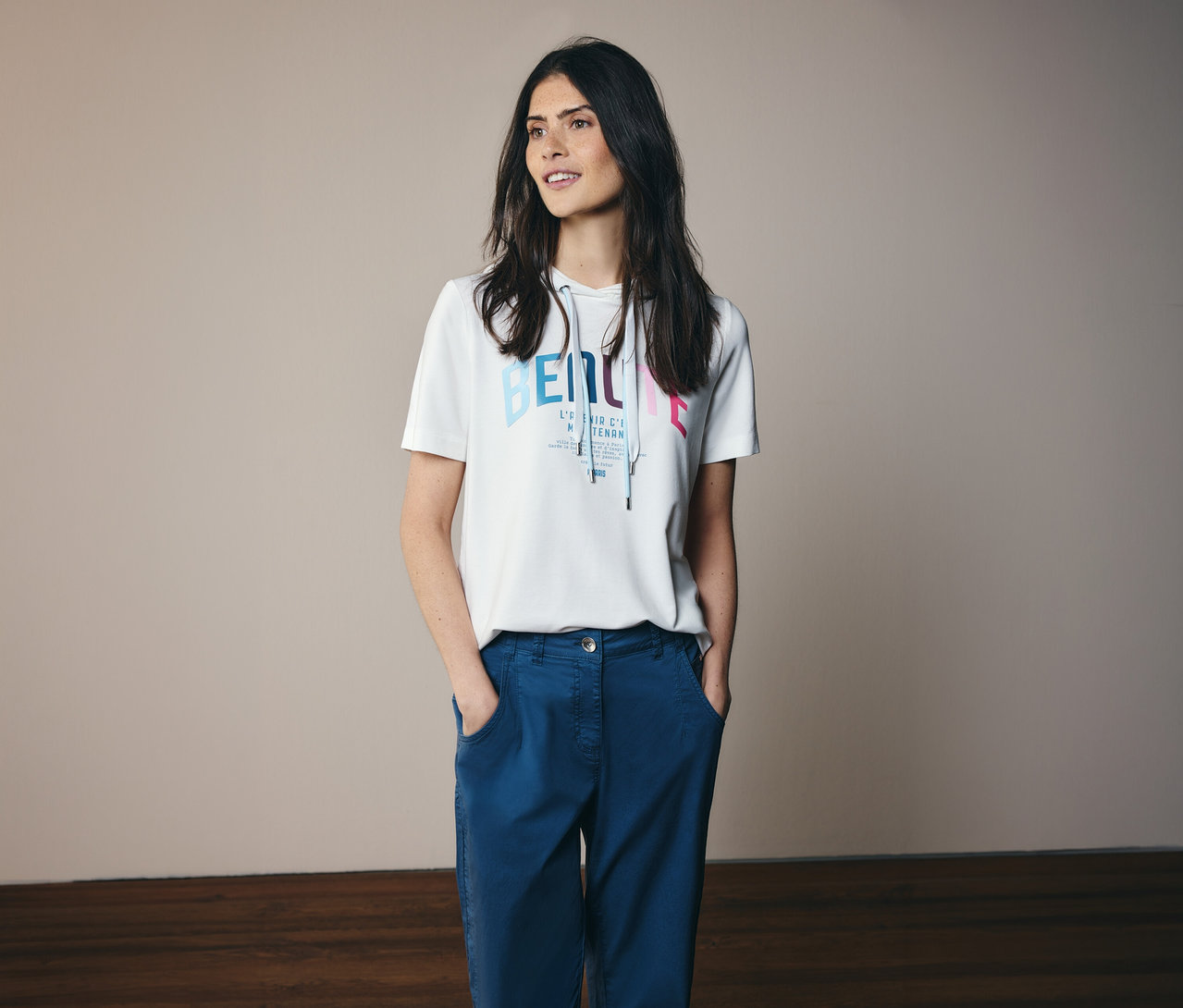 Žena v modrých nohaviciach a bielom mikinovom tričku CECIL s potlačou a kapucňou, s rukami vo vreckách.