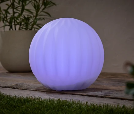 Okrúhla, fialová lampa stojí na drevenom stole vedľa črepníkovej rastliny.