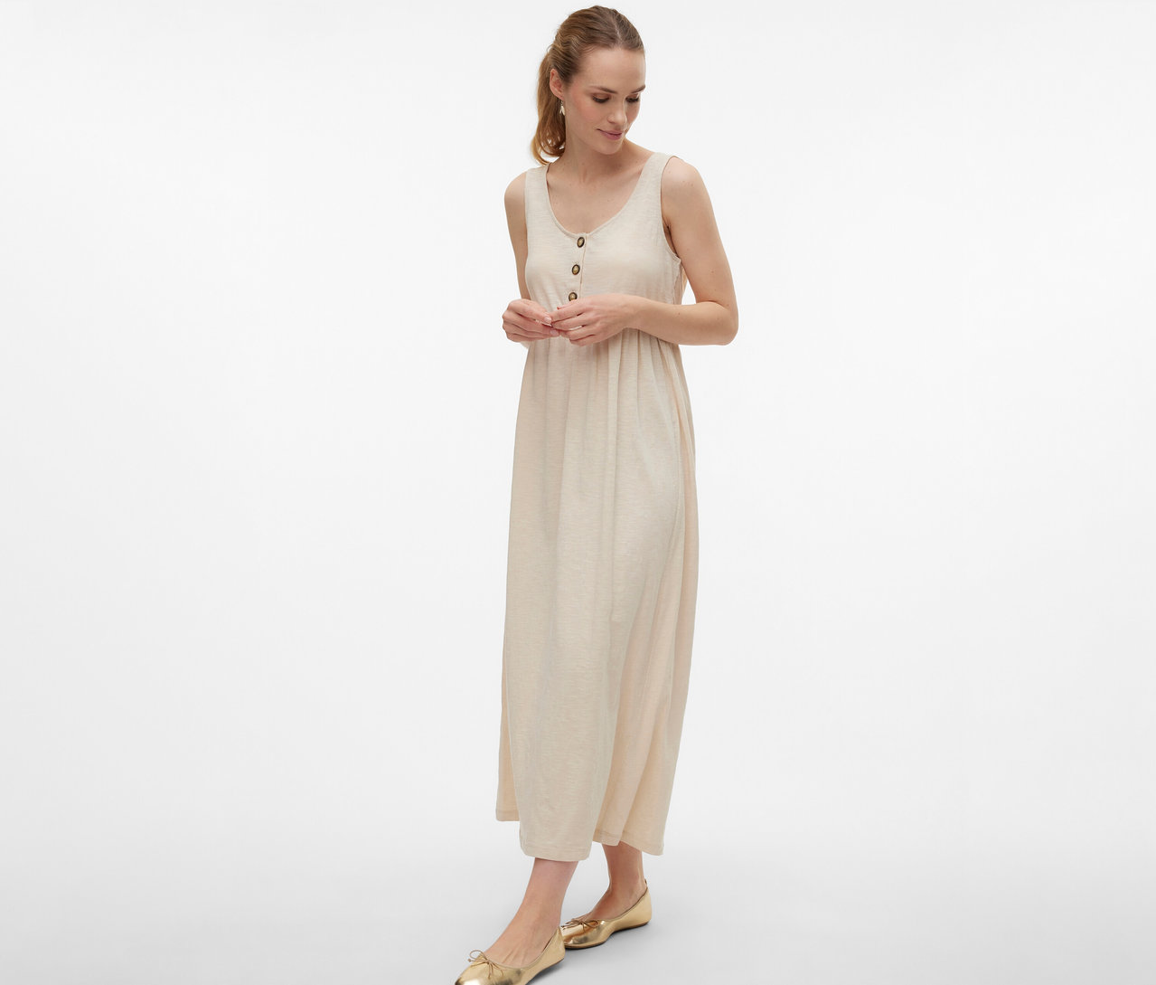 Žena nosí tehotenské šaty MAMALICIOUS »MLEVI LIA« s gombíkmi a zlaté baleríny na bielom pozadí.