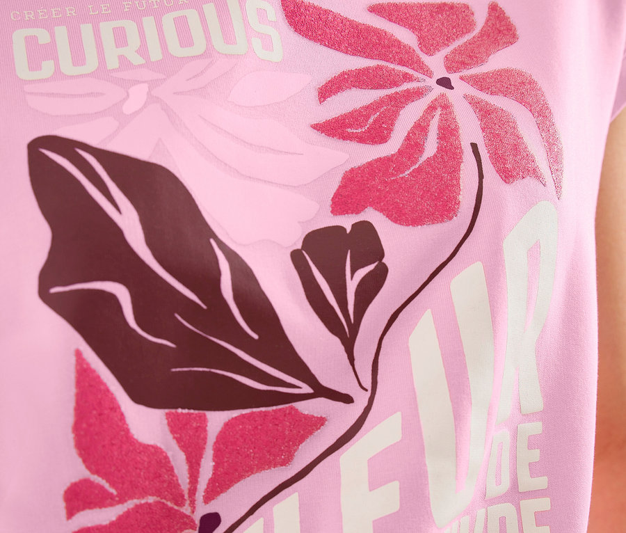 Osoba má na sebe ružové Placement tričko CECIL s kvetinovou potlačou v ružovej a tmavohnedej farbe a nápismi "CURIOUS" a "FLEUR DE VIVRE".