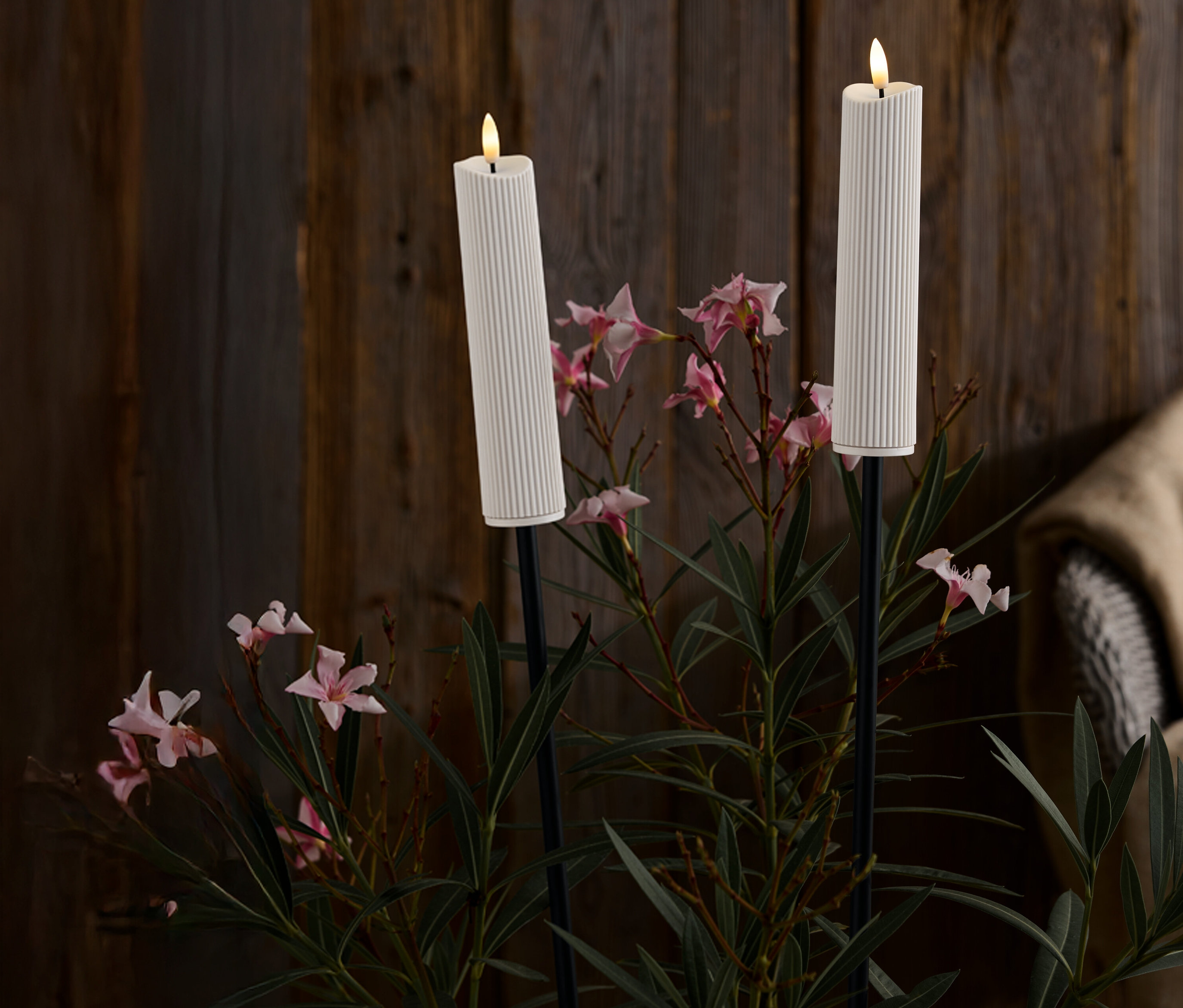 Dve biele, rebrované sviečky na čiernych stojanoch uprostred ružových kvetov a zelených listov na pozadí tmavého dreva.