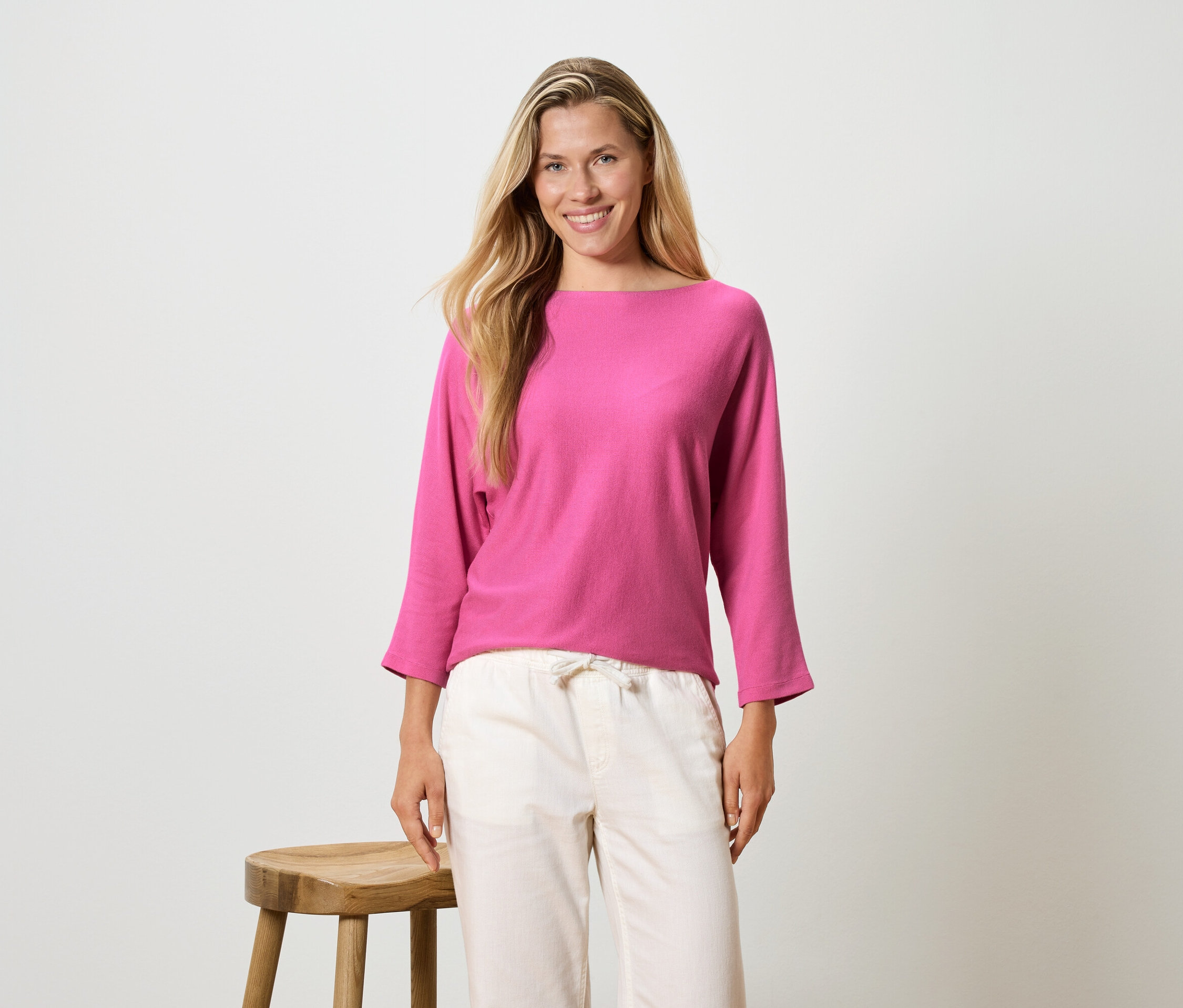 Žena s dlhými blond vlasmi má na sebe ružovú blúzku s dlhým rukávom a svetlé nohavice, stojí vedľa stoličky.