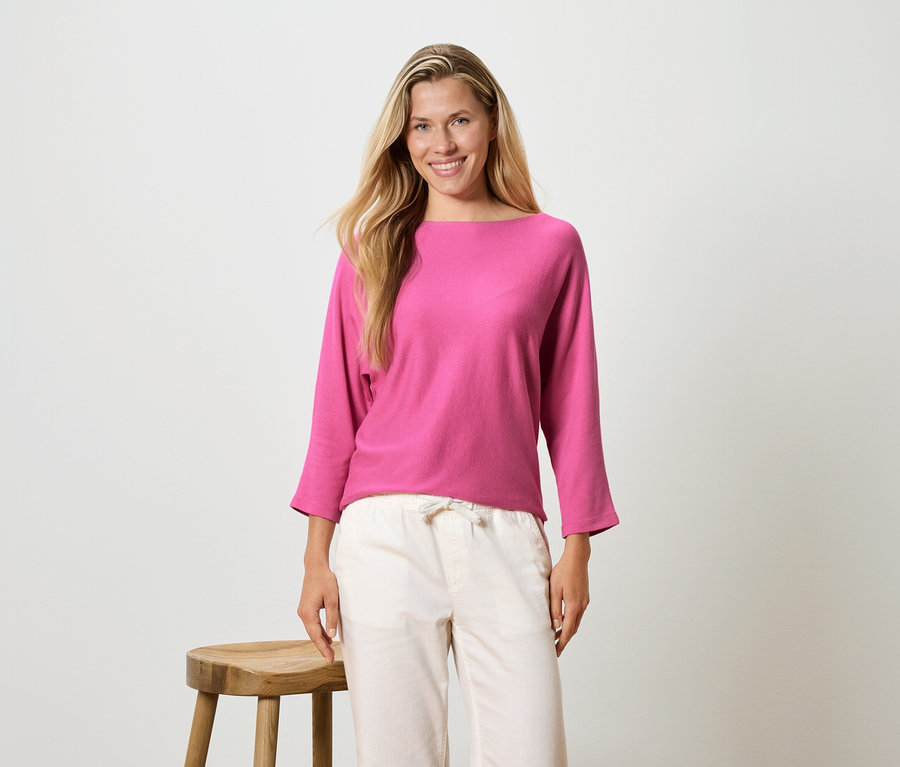Žena s dlhými blond vlasmi má na sebe ružovú blúzku s dlhým rukávom a svetlé nohavice, stojí vedľa stoličky.