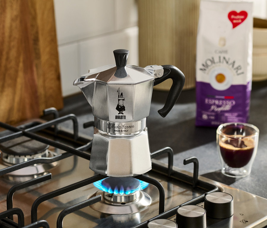 Strieborný espresso kávovar stojí na plynovom sporáku s modrým plameňom, vedľa neho šálka espressa a balenie kávy Molinari.