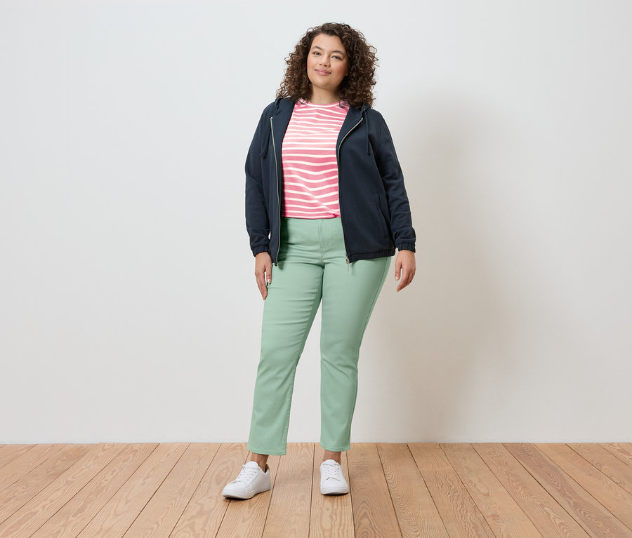Žena stojí v svetlom prostredí a má na sebe tmavomodrú bundu, pruhovaný top, svetlozelené nohavice a biele tenisky.