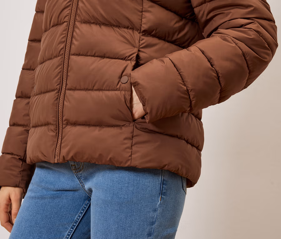 Detail osoby v džínsoch a hnedej prešívanej bunde s rukou vo vrecku.