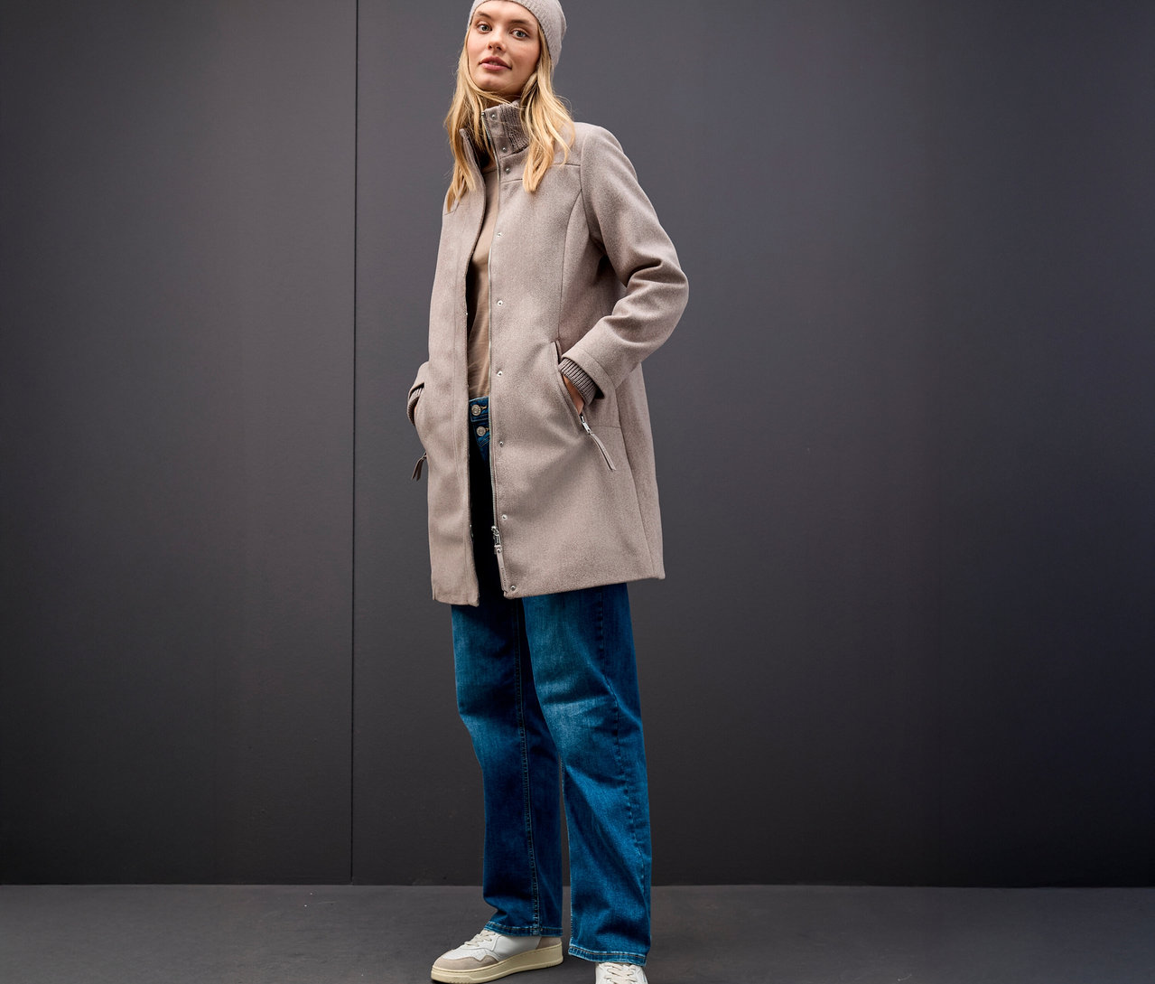 Žena s béžovou čiapkou a krátkym kabátom Street One na sivom pozadí.