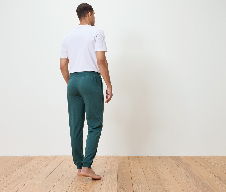 Muž zozadu v bielom tričku a zelených teplákoch, stojaci na drevenej podlahe pred bielou stenou.