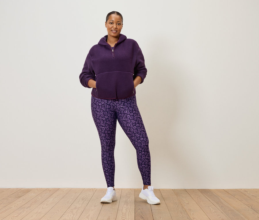 Žena v purpurovej flísovej mikine na polovičný zips a purpurových legínach s leopardím vzorom, ktorá má na sebe biele tenisky.