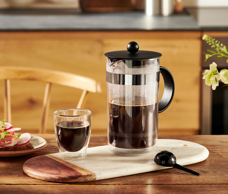 Kanvica french press Bodum® 1 l stojí na stole vedľa šálky kávy a taniera s chlebíčkami.