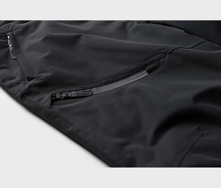 Detail čiernej technickej látky s dvoma vreckami na zips; jedno horizontálne, druhé diagonálne, obe s úchytkami „ACTIVE“.
