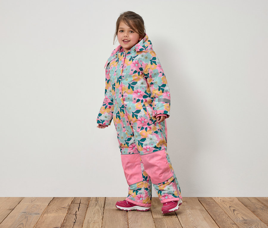 Usmievajúce sa dievčatko v kvetinovom overale s kapucňou a ružovými topánkami stojí na drevenej podlahe.