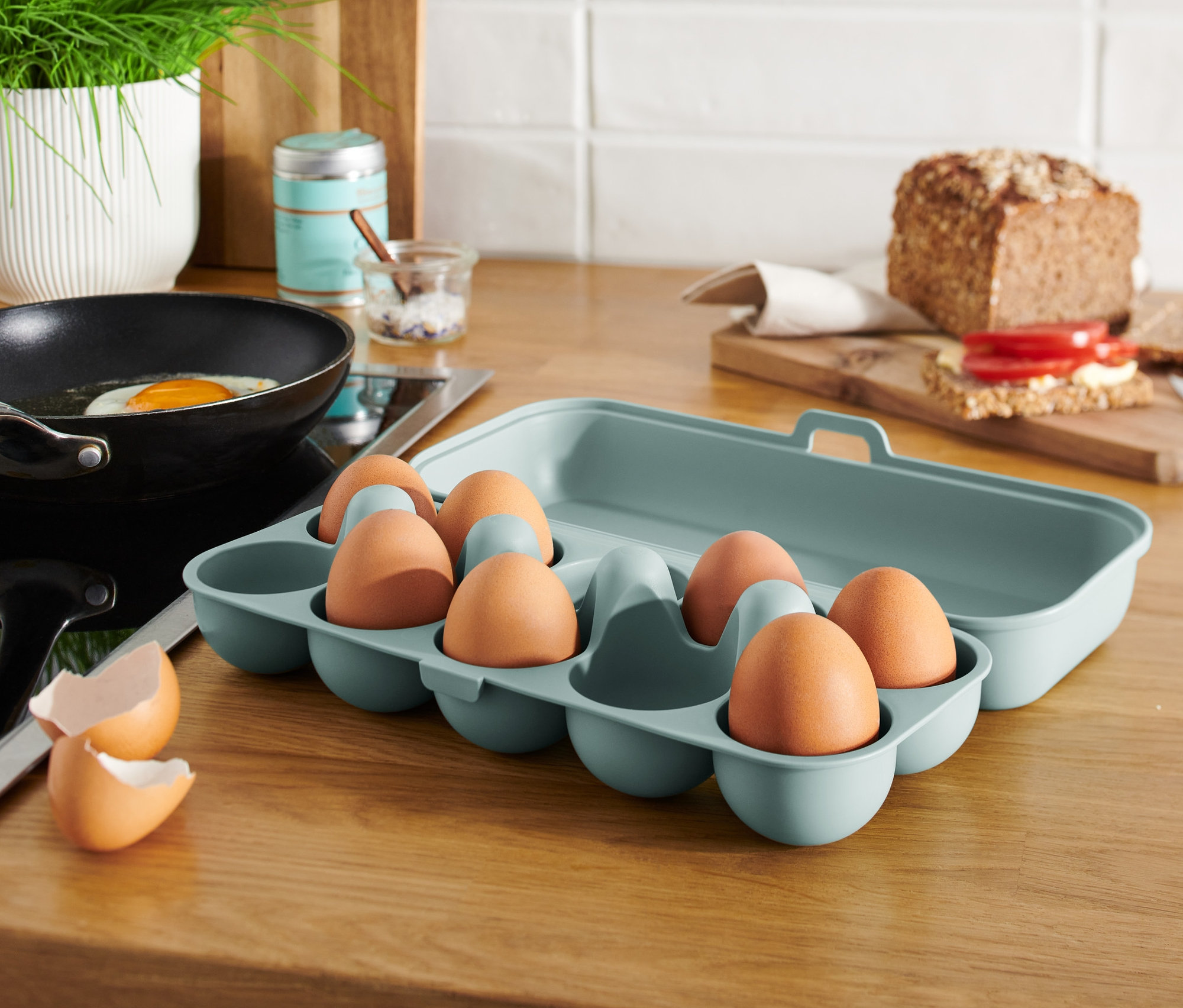 Vajcia v modrozelenej dóze na vajíčka na kuchynskej linke vedľa panvice s vajcom.
