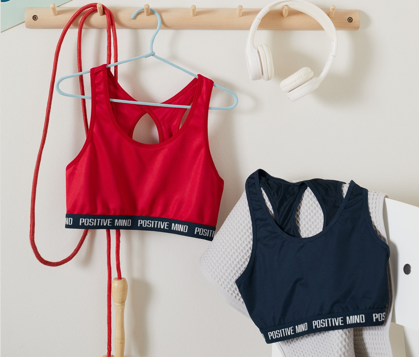 Červená športová podprsenka z 2 ks visí na vešiaku vedľa švihadla. Modrá športová podprsenka z 2 ks leží na sivej stoličke vedľa uteráka.