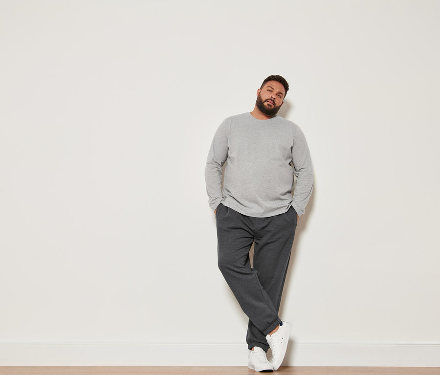 Muž opretý o stenu má na sebe tričko s dlhým rukávom, teplákové nohavice a topánky sneaker.