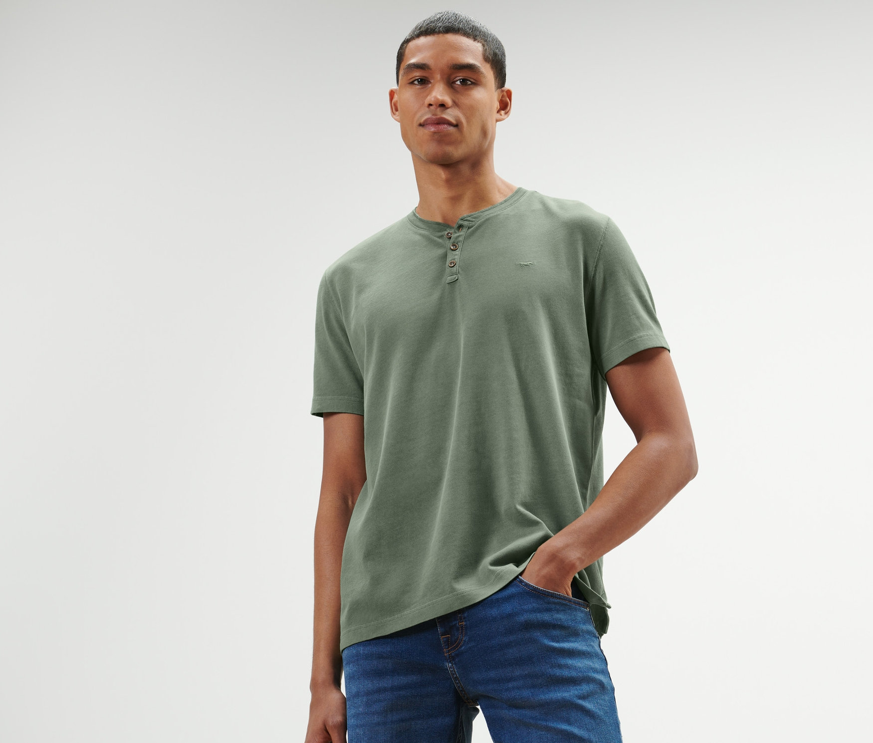 Muž s krátkymi vlasmi pózuje v zelenom tričku henley »Mustang« a modrých džínsoch.