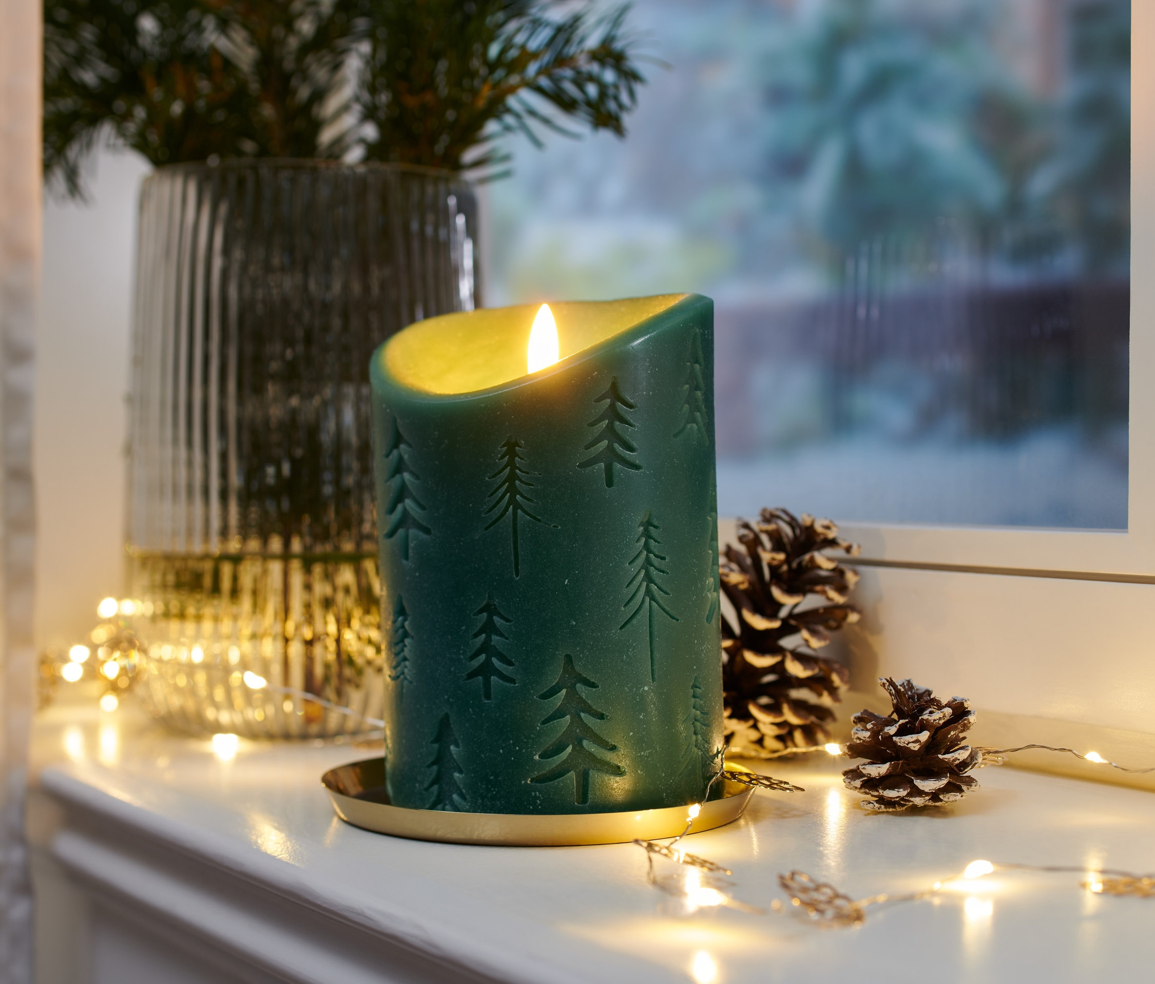 Zelená LED sviečka z pravého vosku so vzorom vianočného stromčeka na zlatom podnose so šiškami a svetielkami.