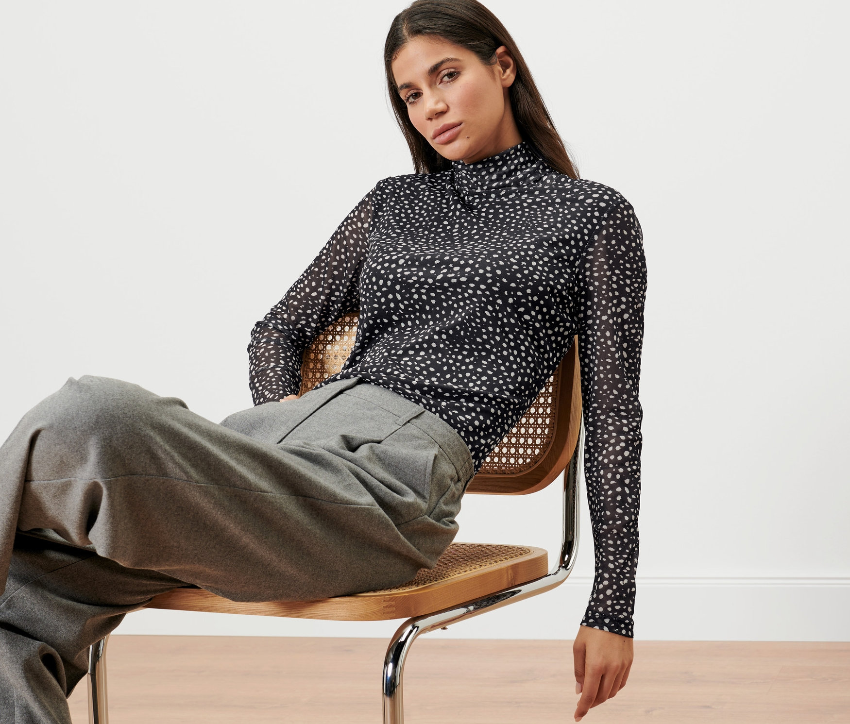 Žena sedí na stoličke a má na sebe bodkované tričko zo sieťoviny s rolákovým golierom.