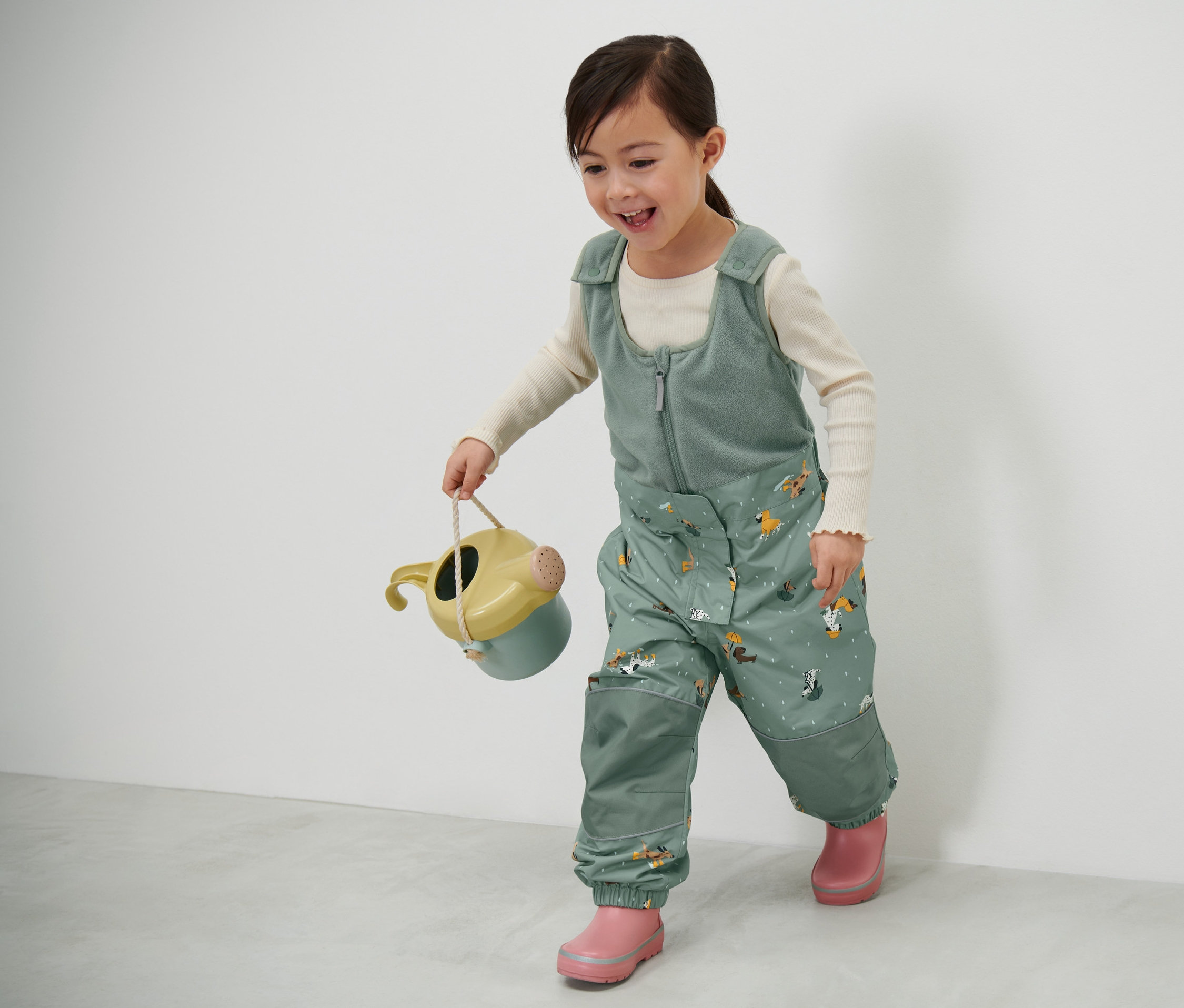 Dievčatko nesie kanvicu a má na sebe detské termonohavice do dažďa.