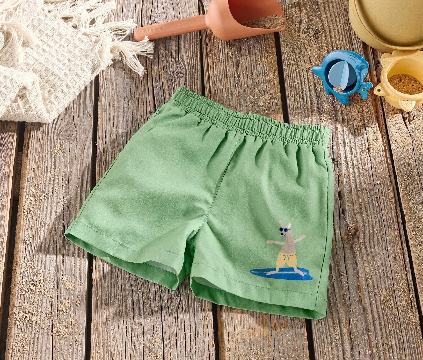 Zelené detské board šortky s obrázkom kengury na surfovej doske ležia na drevenom stole s hračkami do piesku.