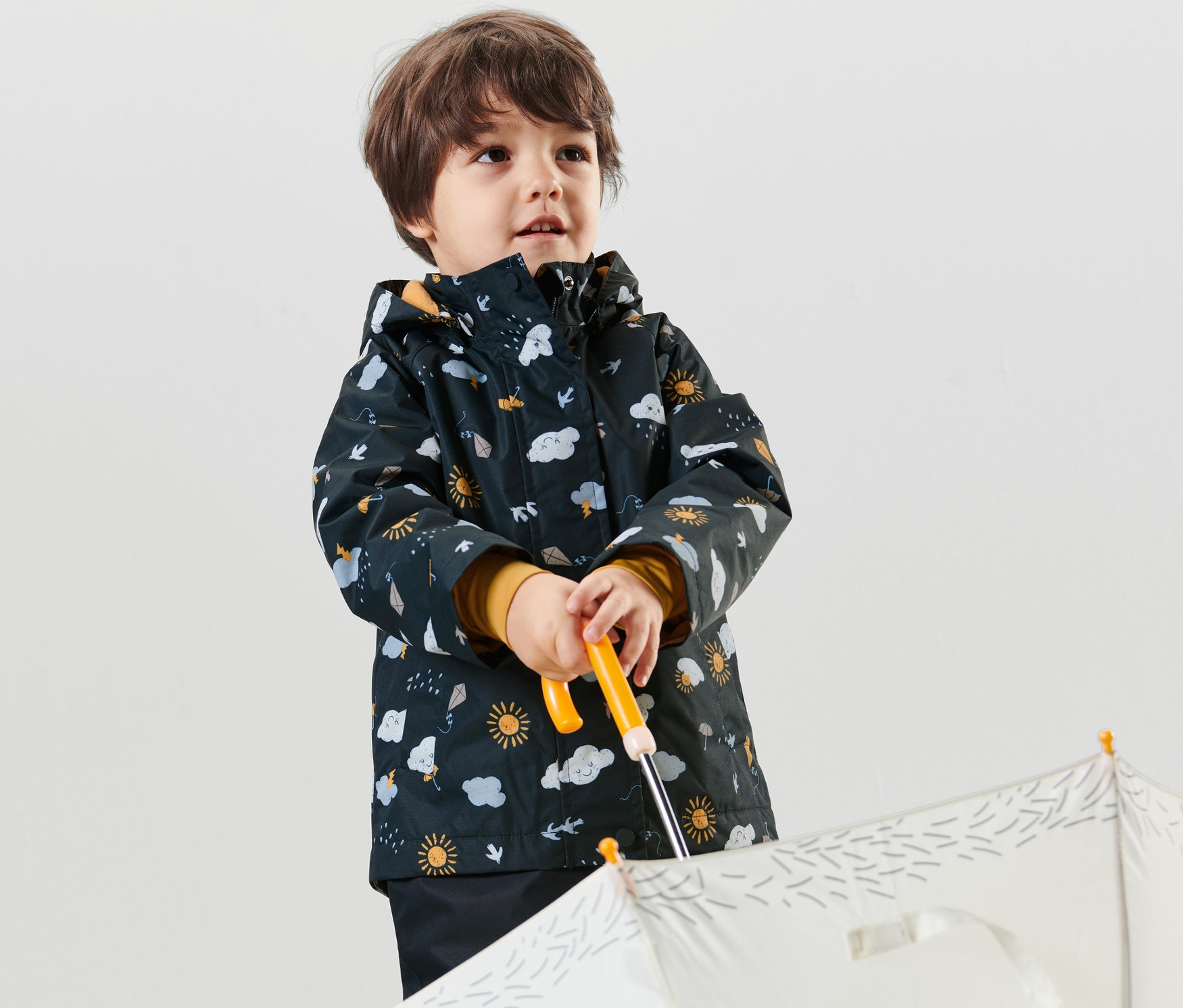 Chlapec drží biely dáždnik a má na sebe tmavomodrú detskú termo bundu do dažďa s celoplošnou potlačou.