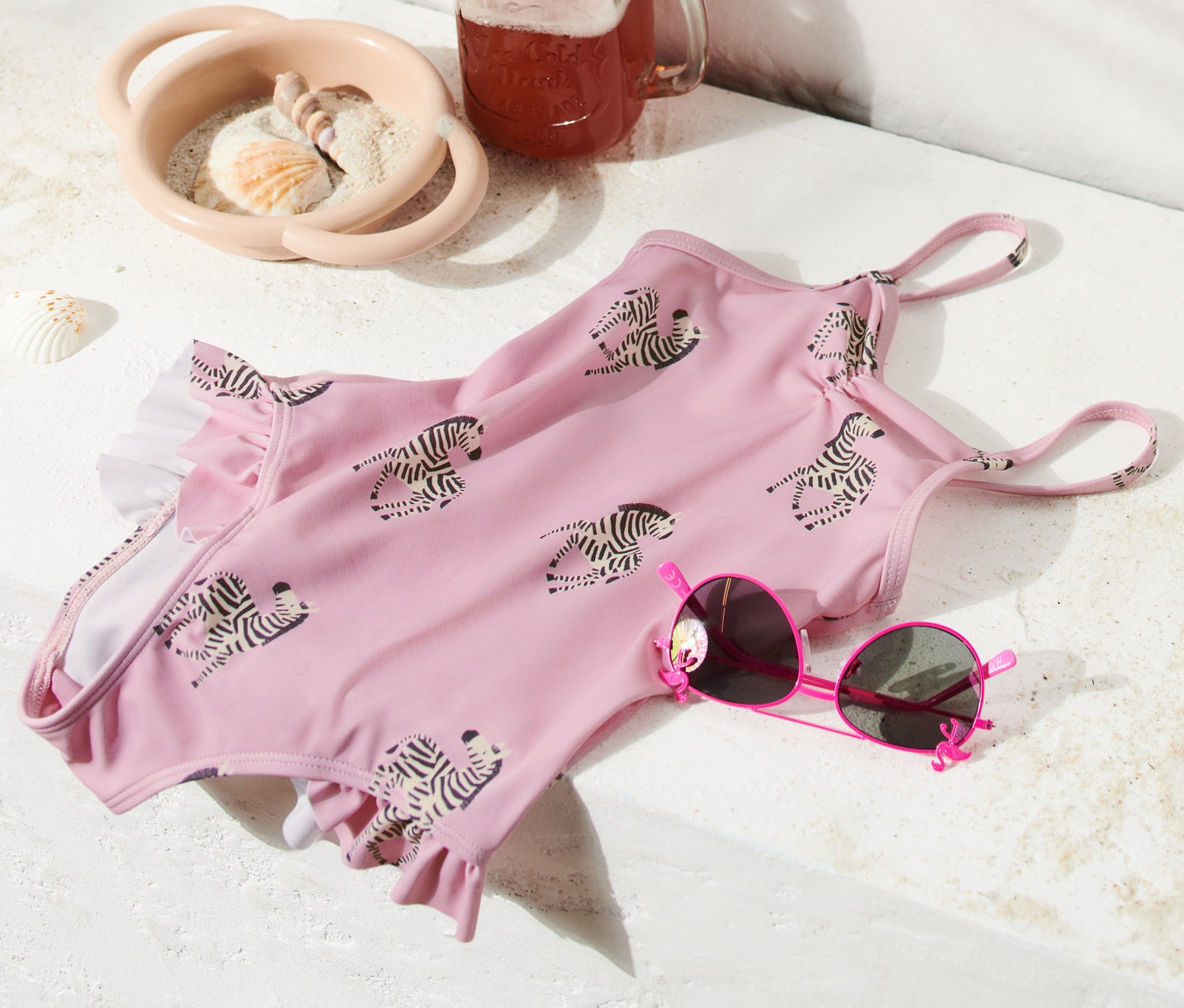 Ružové jednodielne plavky s volánikmi so vzorom zebry ležia vedľa ružových slnečných okuliarov, pohára s nápojom a plážovej tašky.