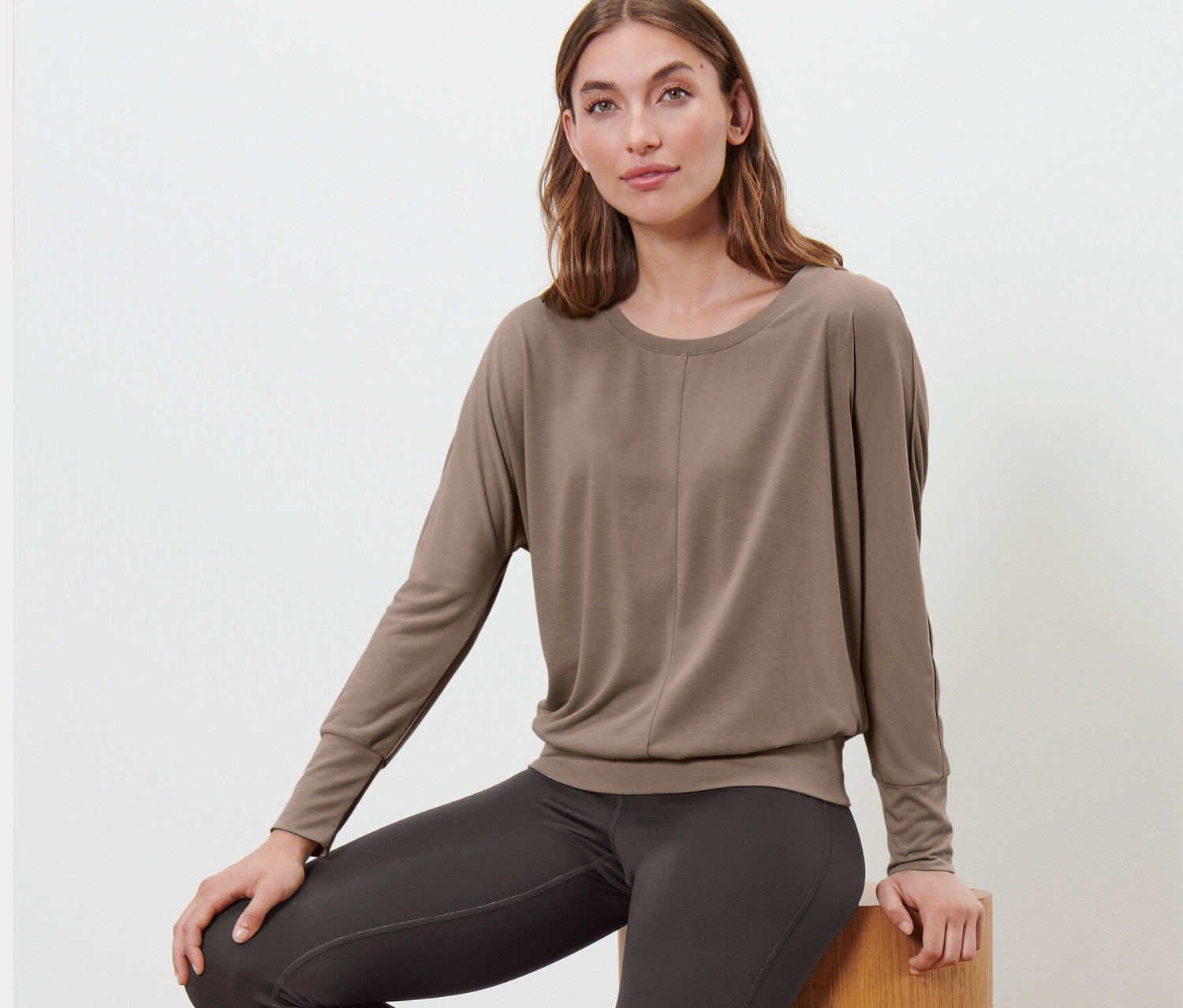 Žena sedí na drevených stupňoch a má na sebe hnedosivé tričko na šport a jogu.