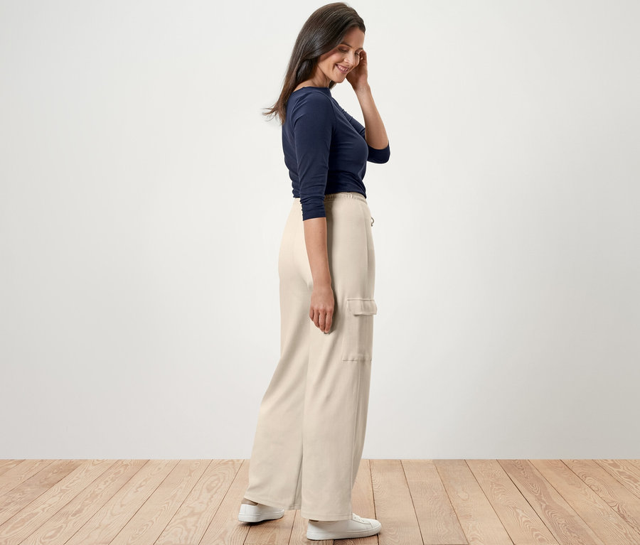 Žena stojí bokom a má na sebe krémovobiele teplákové nohavice s cargo vreckami, modré tričko a biele tenisky.