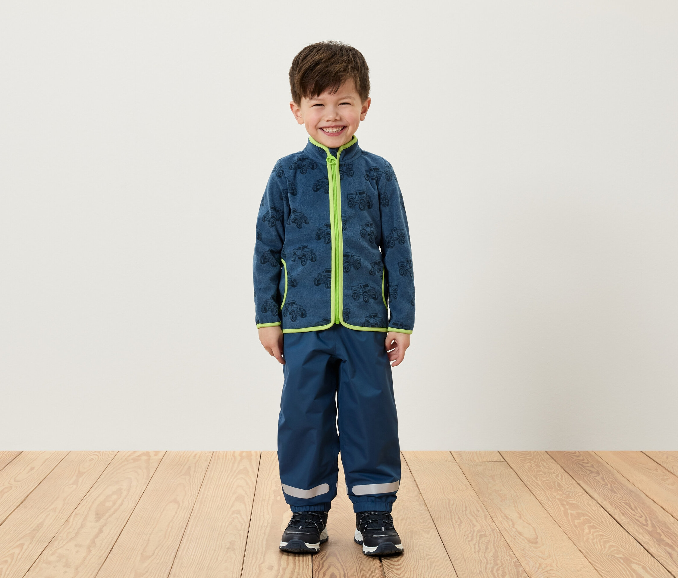 Chlapec stojí a má na sebe modrú flísovú bundu so zeleným lemovaním a modré nohavice.