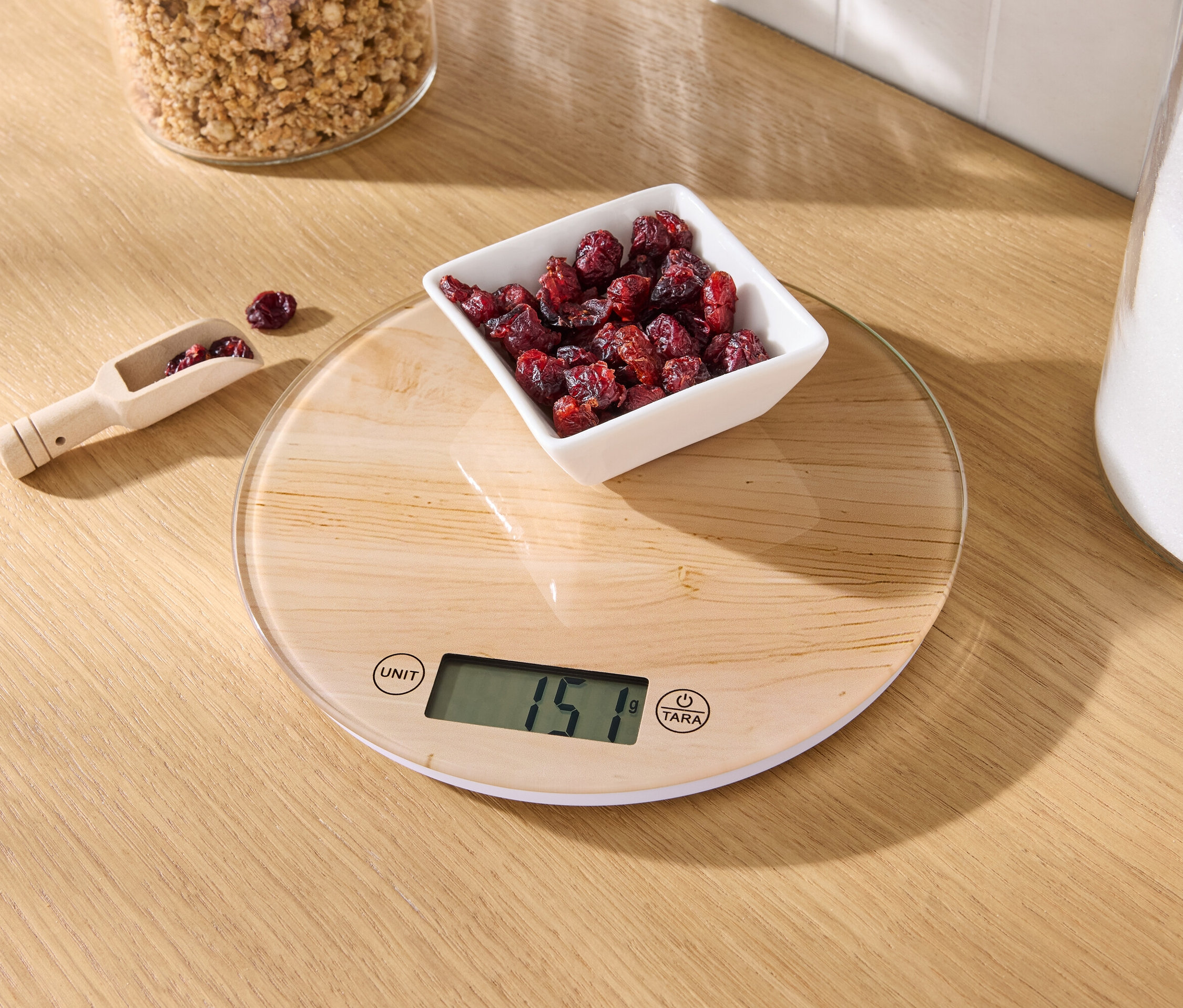 Na drevenom stole stojí kuchynská váha s miskou sušených brusníc a drevenou odmerkou s brusnicami.