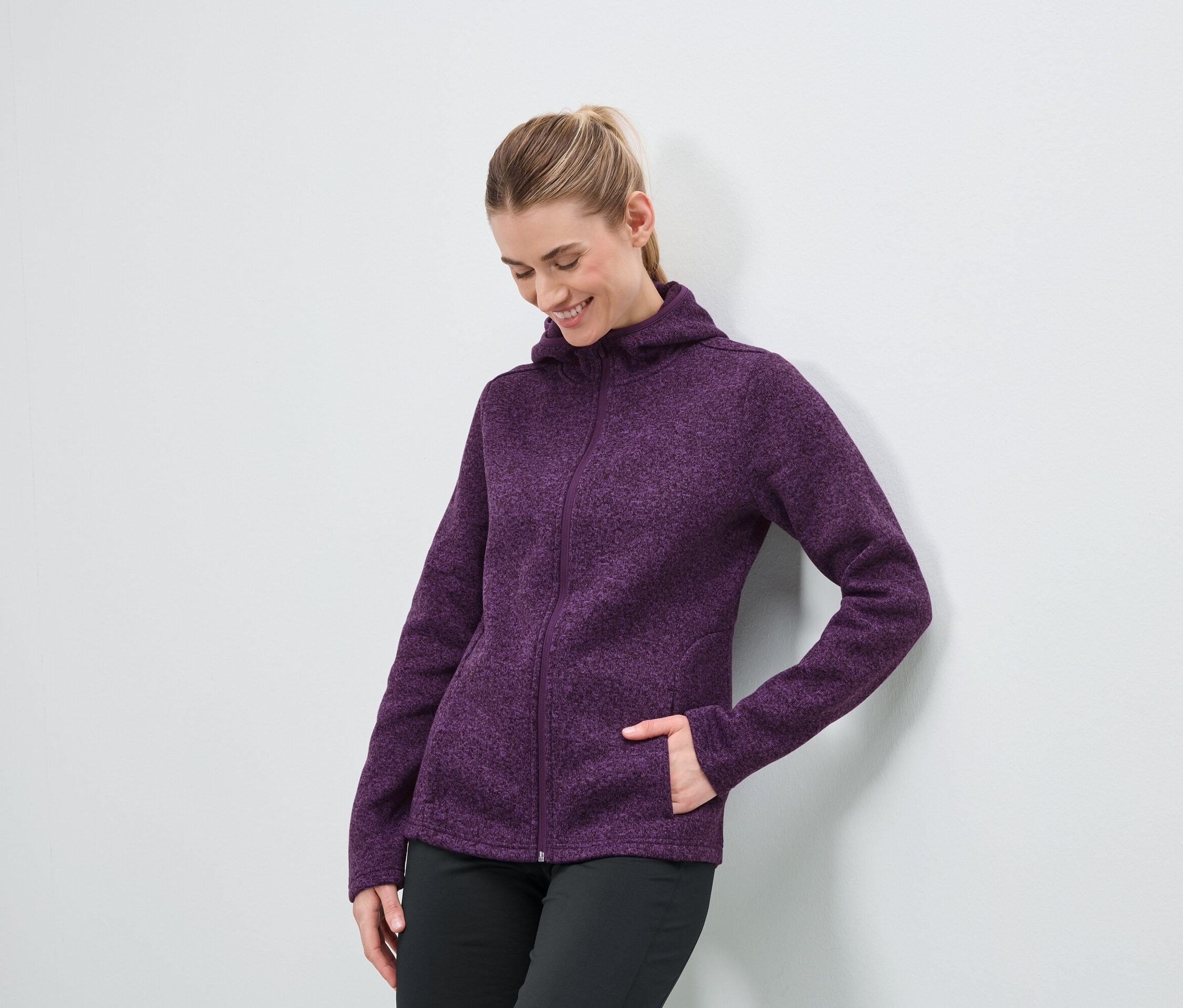 Žena v purpurovej mikine s kapucňou na zips a čiernych nohaviciach stojí pri bielej stene.