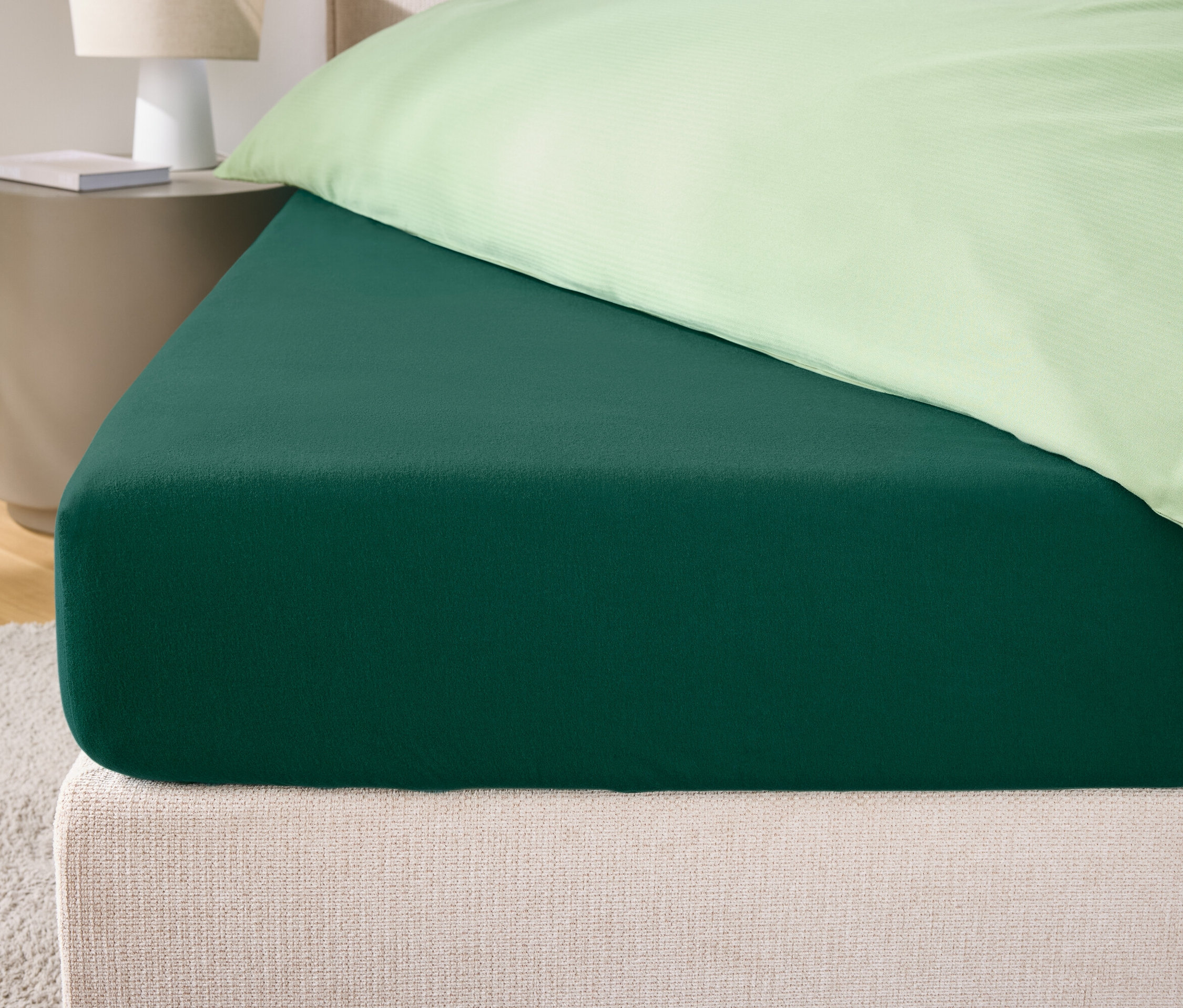 Detail zelenej napínacej plachty na posteli s krémovým čelom a svetlozelenou obliečkou na vankúš.