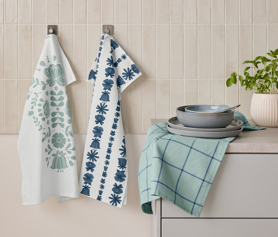 Dve utierky s kvetinovými vzormi visia vedľa seba na stene. Pod nimi leží zložená zelená utierka s modrými pruhmi a na nej naskladané taniere a miska. Vedľa stojí kvetináč s bylinkami.