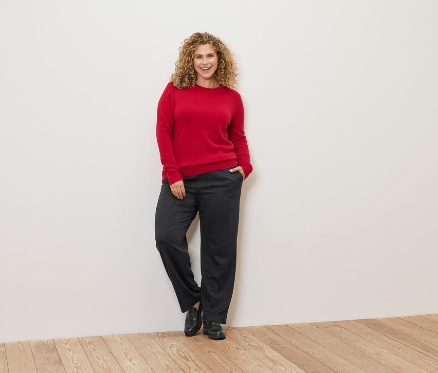 Žena s kučeravými vlasmi sa opiera o stenu a má na sebe červený sveter a tmavé nohavice.