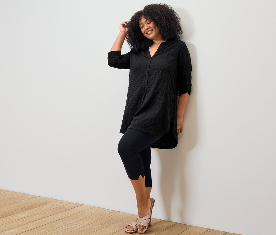 Žena pózuje v čiernej tunike s tkanou štruktúrou a legínach s detailom v 3/4 dĺžke.