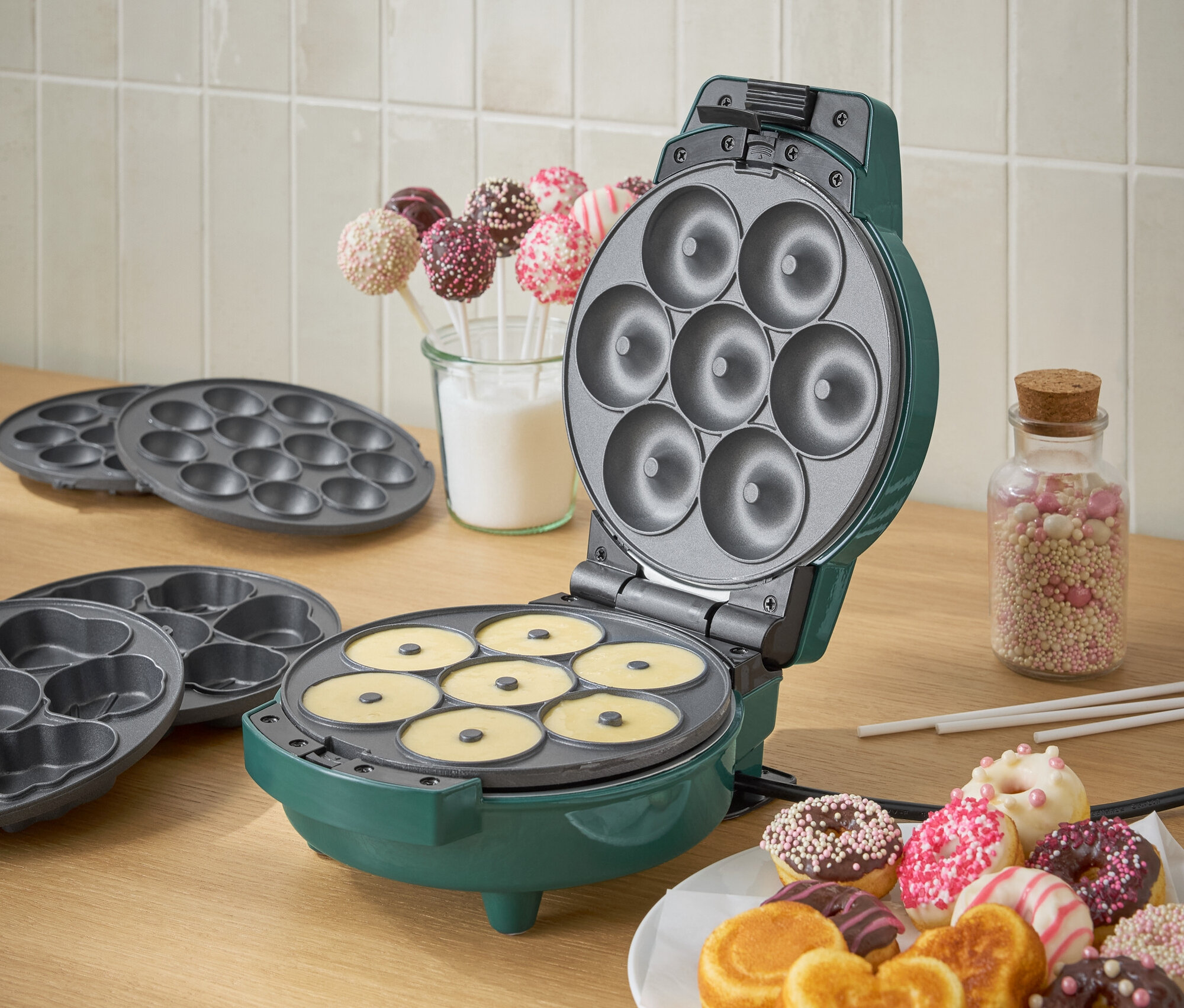 Zelený výrobník na donuty s cestom a tanierom hotových donutov.