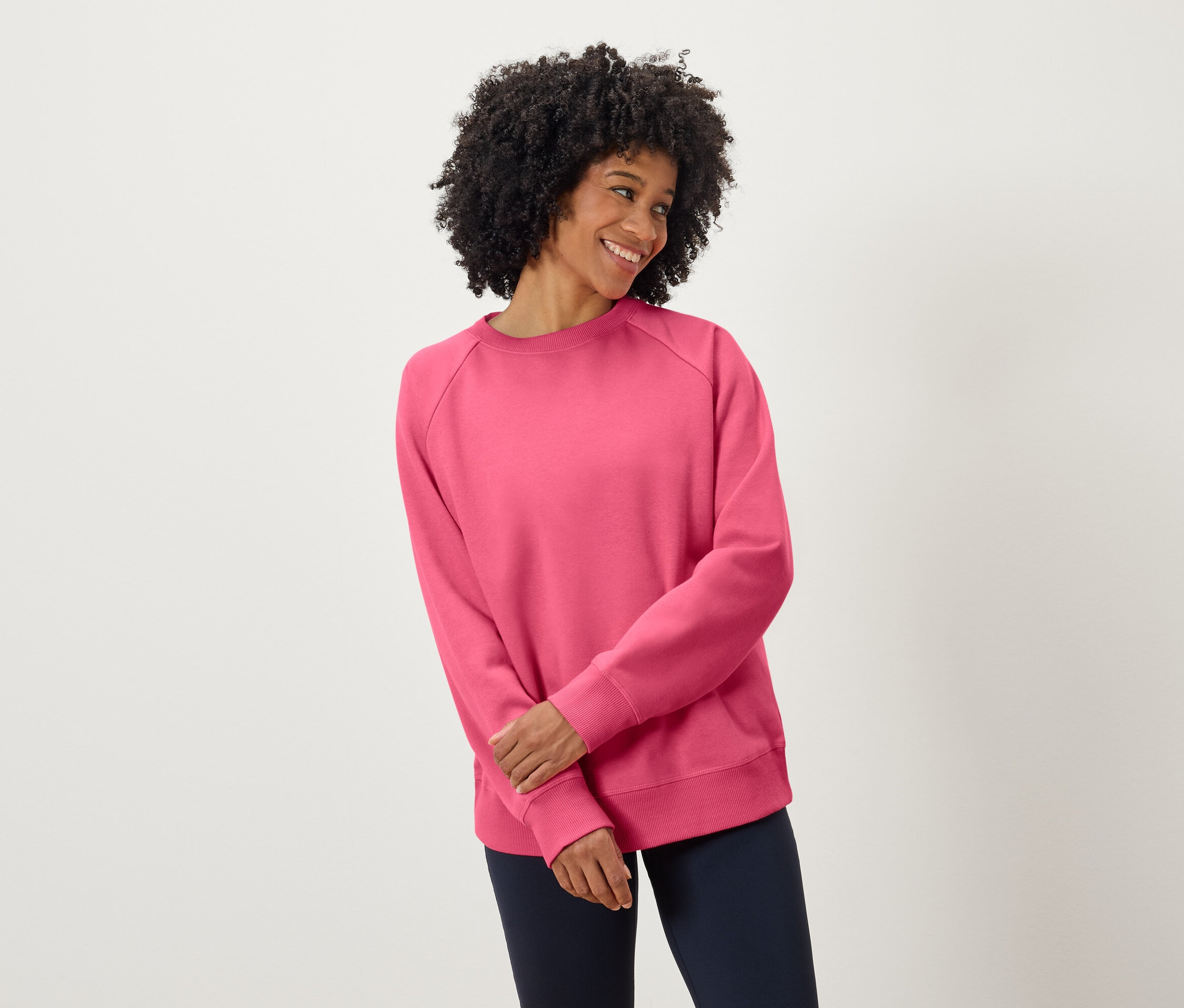 Žena s tmavými kučeravými vlasmi sa usmieva a má na sebe ružovú mikinu a tmavomodré nohavice.