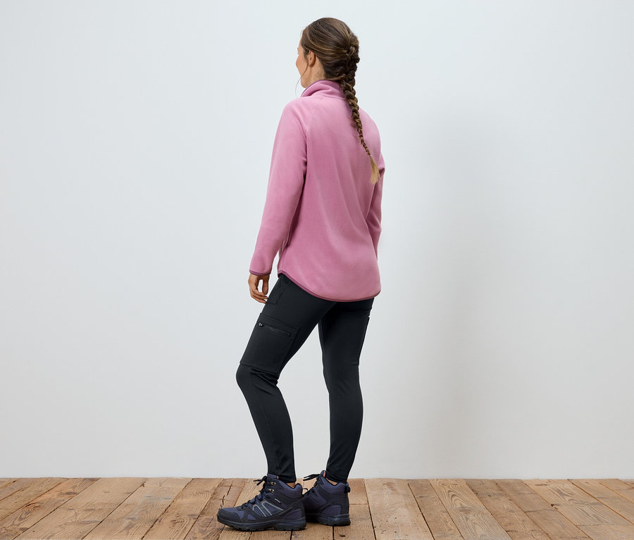 Žena v ružovej flísovej bunde, čiernych nohaviciach a topánkach stojí chrbtom k fotoaparátu.