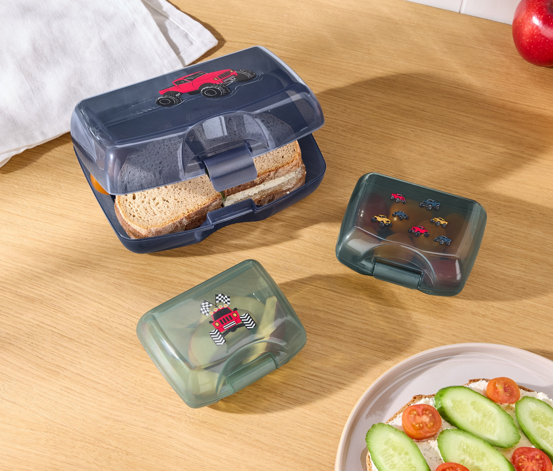Na drevenom stole leží otvorený modrý box na desiatu s obrázkom červeného auta, dva zelené boxy na desiatu s obrázkami áut, jablko, pomaranč, biela utierka a tanier s chlebíčkom.