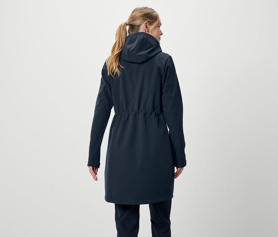 Zozadu je vidieť žena s blond vlasmi stiahnutými do copu, má na sebe tmavomodrý kabát a nohavice.